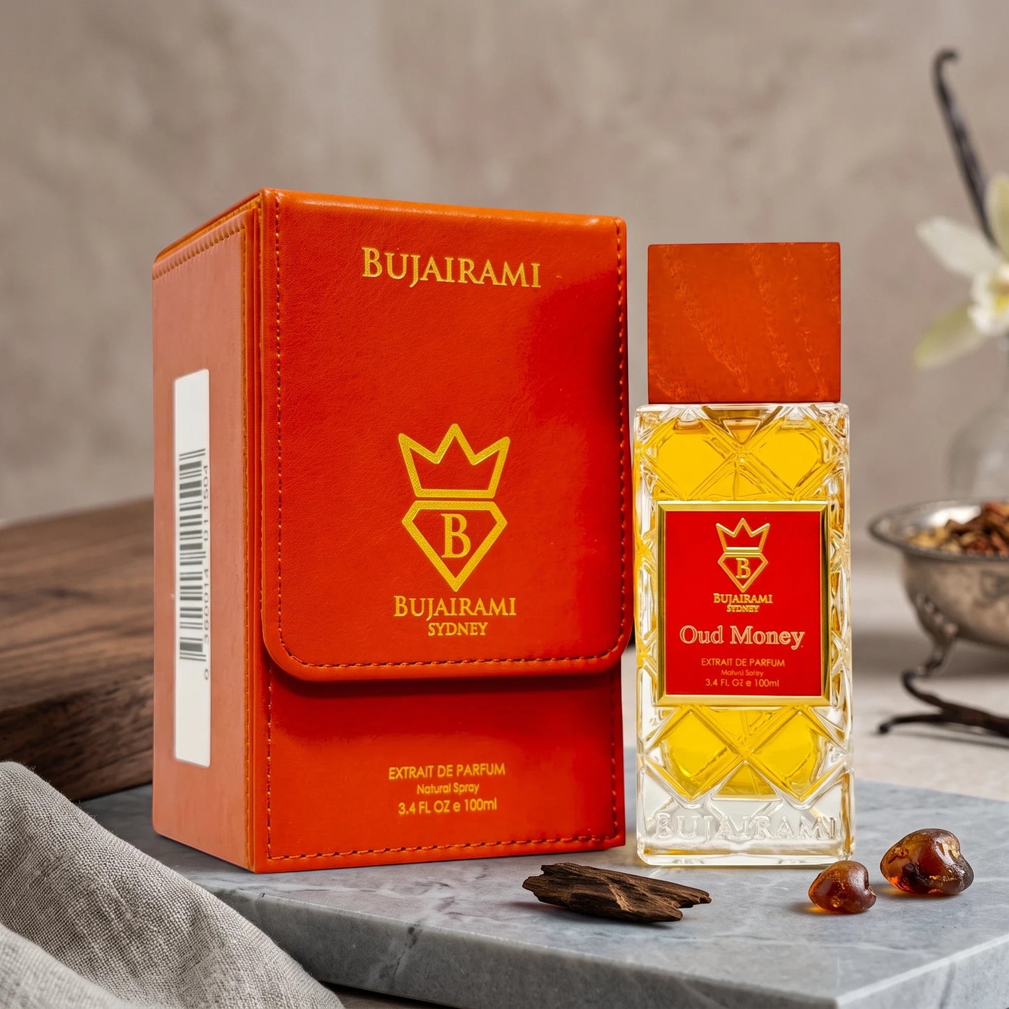Soghaat Bujairami Oud Money Extrait de Parfum bottle and orange leather case on a stone surface with oud wood chips.