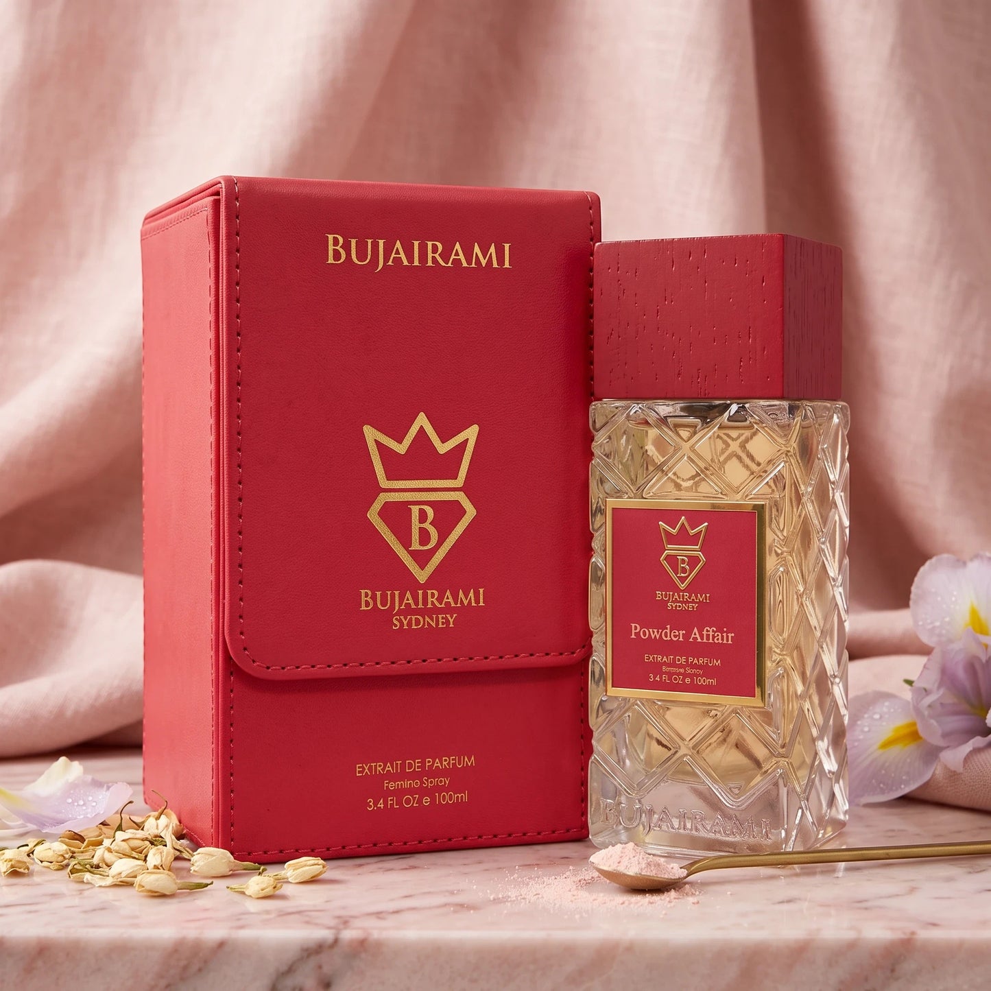 Soghaat Bujairami Powder Affair Extrait de Parfum bottle and red leather case on a marble surface with flower petals.