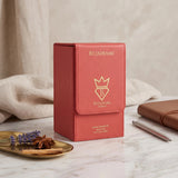 Soghaat Bujairami Powder Affair 100ml Extrait de Parfum in a coral-coloured leather-textured box on a marble surface.