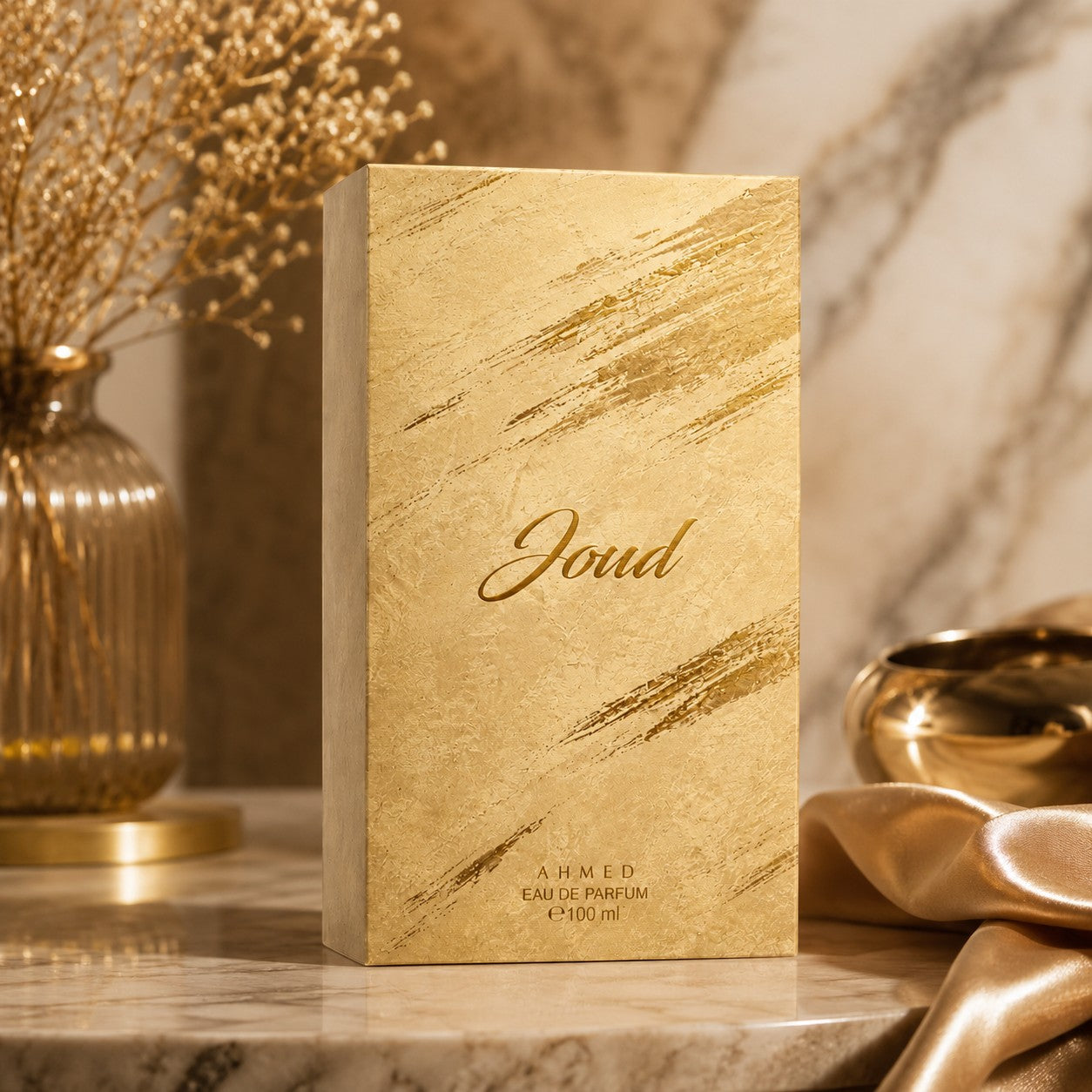 Soghaat Joud Eau de Parfum by Ahmed Al Maghribi in a textured gold box on a marble surface with silk and dried flowers.