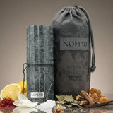 Soghaat Nomad Extrait de Parfum bottle with grey textured packaging and a drawstring bag. Surrounded by saffron, sea salt, lemon, vanilla, and leaves.