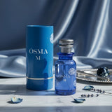 Soghaat Osma M Eau de Parfum bottle and cylindrical box in blue with white floral accents. Sweet gourmand fragrance.